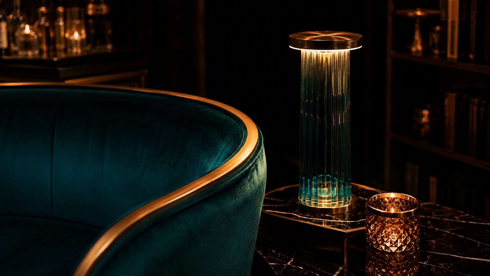 A teal velvet lounge chair beside a glowing glass column lamp and marble side table