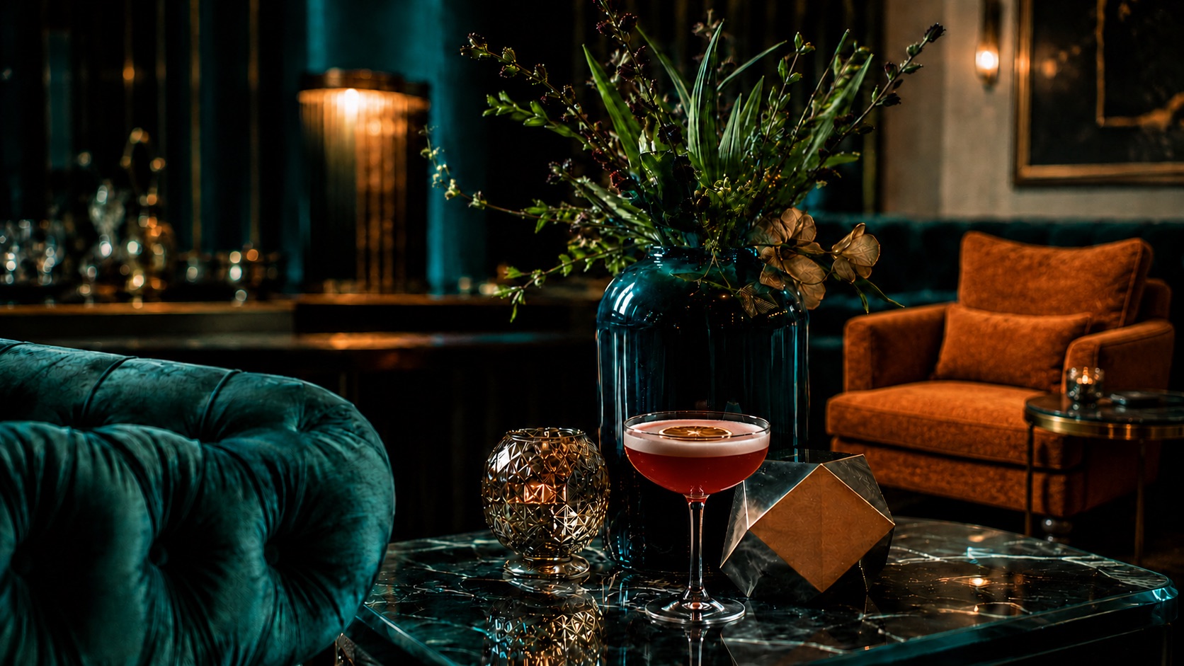 A coupe cocktail with a dried citrus wheel on a black marble table beside a gold votive and blue glass vase, with teal velvet and rust-orange seating